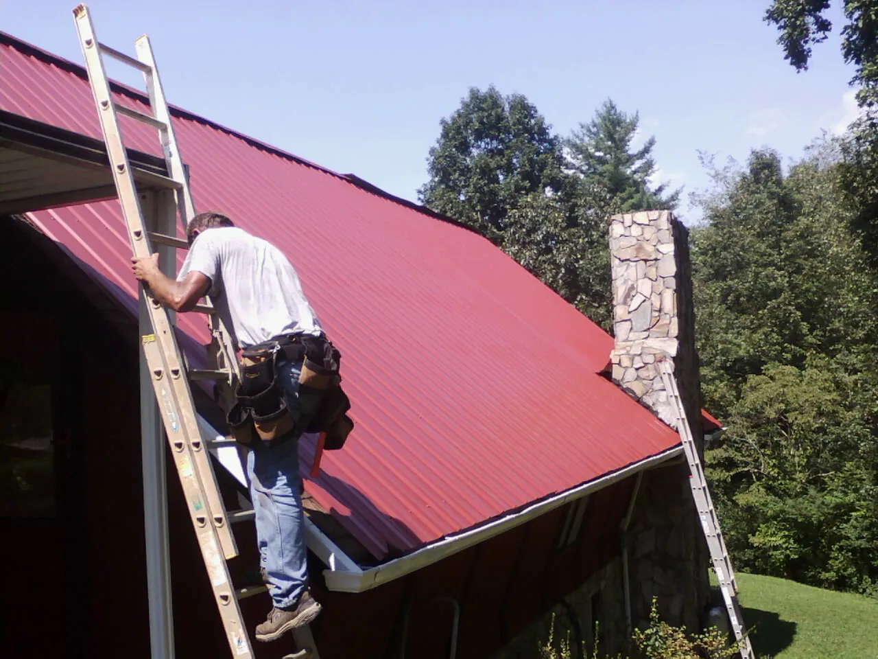 Roofer on a ladder beside a red metal roof and stone chimney — Soffit & Fascia Repair by Roofing Brothers in Westmoreland